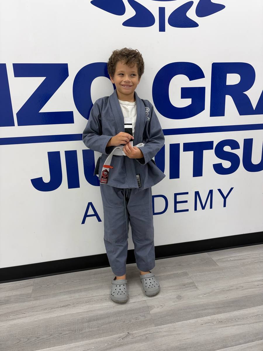 Young student in a grey gi smiling in front of the Renzo Gracie Jiu Jitsu Academy sign